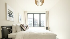 A modern bedroom with a large bed, a bedside table, a lamp, and a view of buildings through a large window. - Chez Meredith (Brussels)