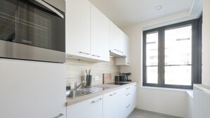 A modern kitchen with white cabinets, a stainless steel oven, a window with blinds, and a view of buildings outside. - Chez Meredith (Brussels)