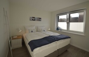 Une chambre à coucher avec un lit, une table de chevet, une lampe de chevet, une fenêtre et un radiateur.