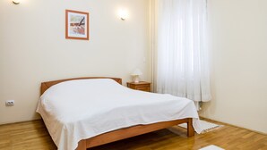 Quarto com cama de madeira, mesa de cabeceira com abajur, quadro na parede e janela com cortinas translúcidas.