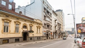 Vista da rua com edifícios, calçada e carros estacionados.