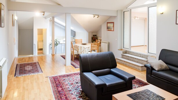 Uma sala de estar ampla com um sofá de couro preto, uma mesa de centro de madeira e uma mesa de jantar com cadeiras. Existe uma escada que leva a um andar superior.