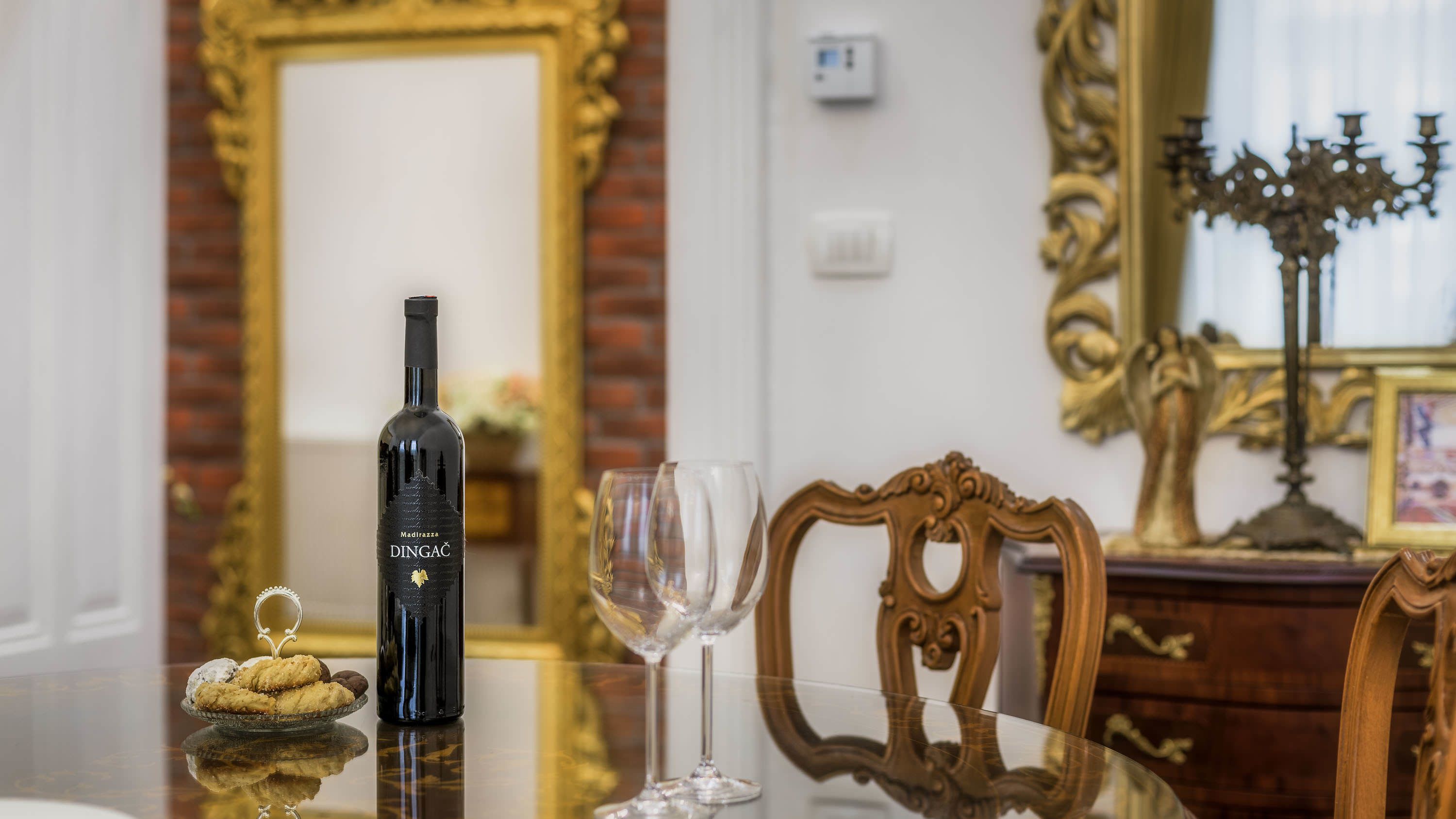 A bottle of Madirazza DINGAČ wine, a glass, and a plate with cookies on a reflective table in a room with ornate furniture and a mirror.