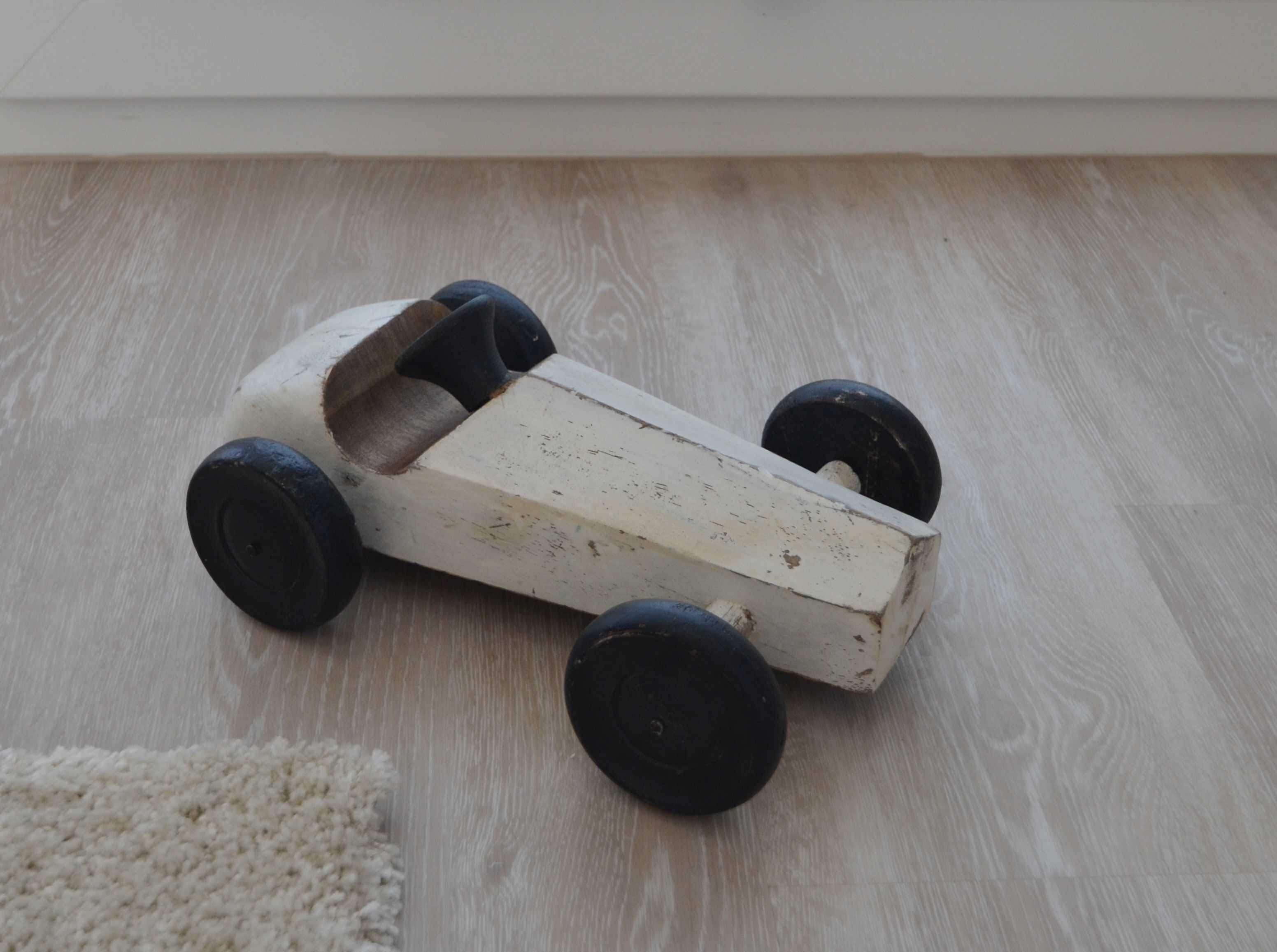 Une petite voiture jouet en bois, blanche, avec des roues noires, posée sur un plancher en bois.