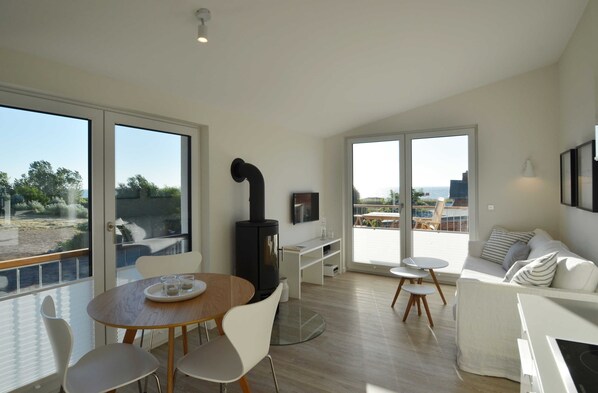 A modern living room with a fireplace, a dining area, and large windows offering a view of the outdoors.