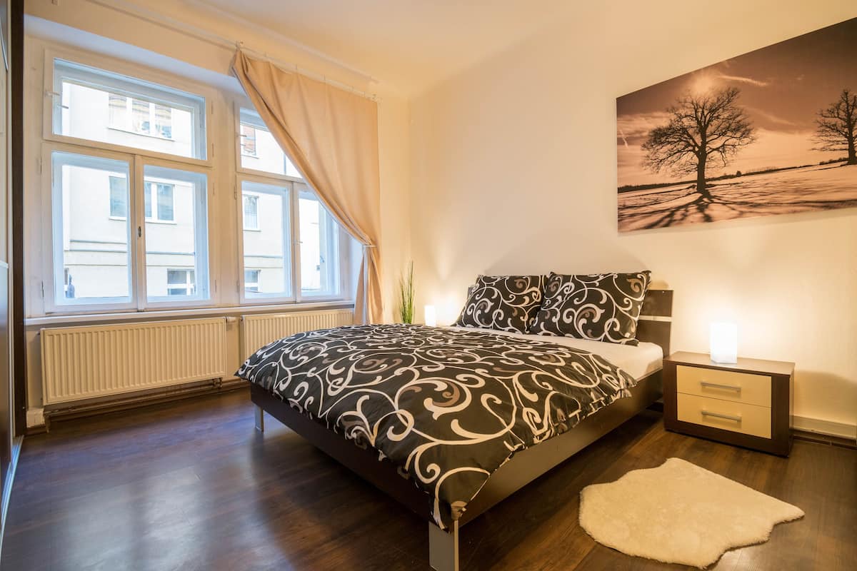 Ein Schlafzimmer mit einem Bett, einem Nachttisch, einer Lampe, einem Fenster mit Vorhängen und einem Wandbild.
