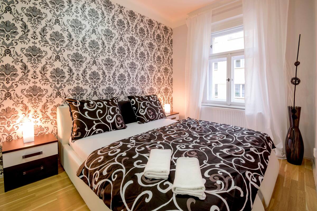Ein ordentlich bezogenes Bett mit einem gemusterten Überwurf, zwei Nachttischlampen, ein Nachttisch mit Schublade und ein Fenster mit Vorhängen.