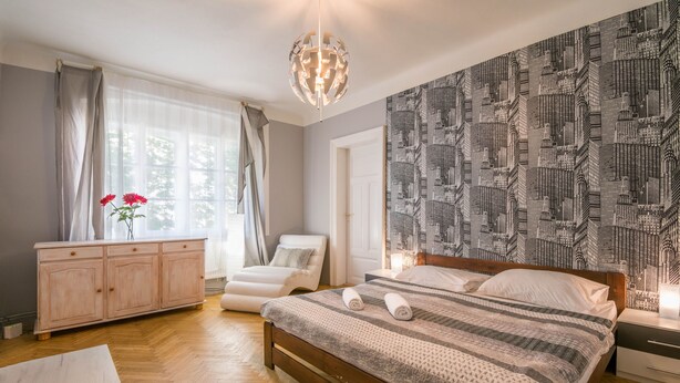 Une chambre à coucher avec un lit en bois, une table de chevet blanche, une chaise blanche, une commode en bois et une grande fenêtre ornée de rideaux légers.
