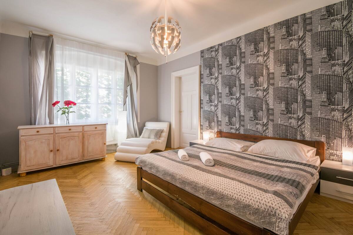 Ein Schlafzimmer mit einem hölzernen Bett, einem weißen Nachttisch, einem weißen Stuhl, einer hölzernen Kommode und einem großen Fenster mit durchscheinenden Vorhängen.