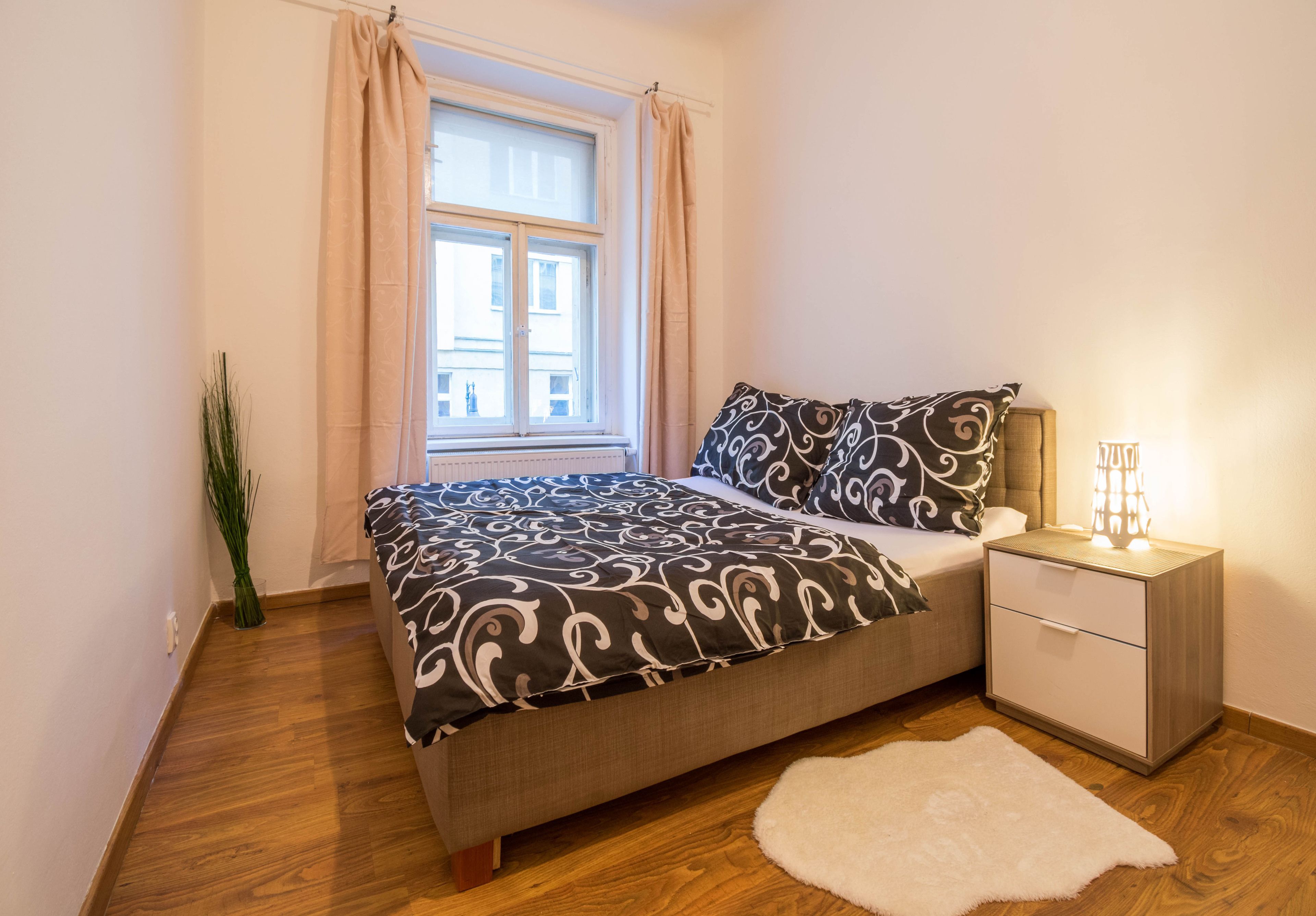 A bedroom with a bed, nightstand, lamp, window with curtains, and a potted plant.