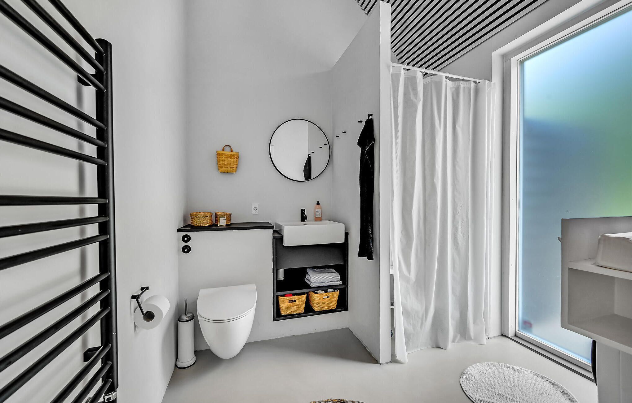 Ein modernes Badezimmer mit Toilette, Waschbecken und Spiegel. Regale mit Körben und ein Fenster mit Vorhängen.