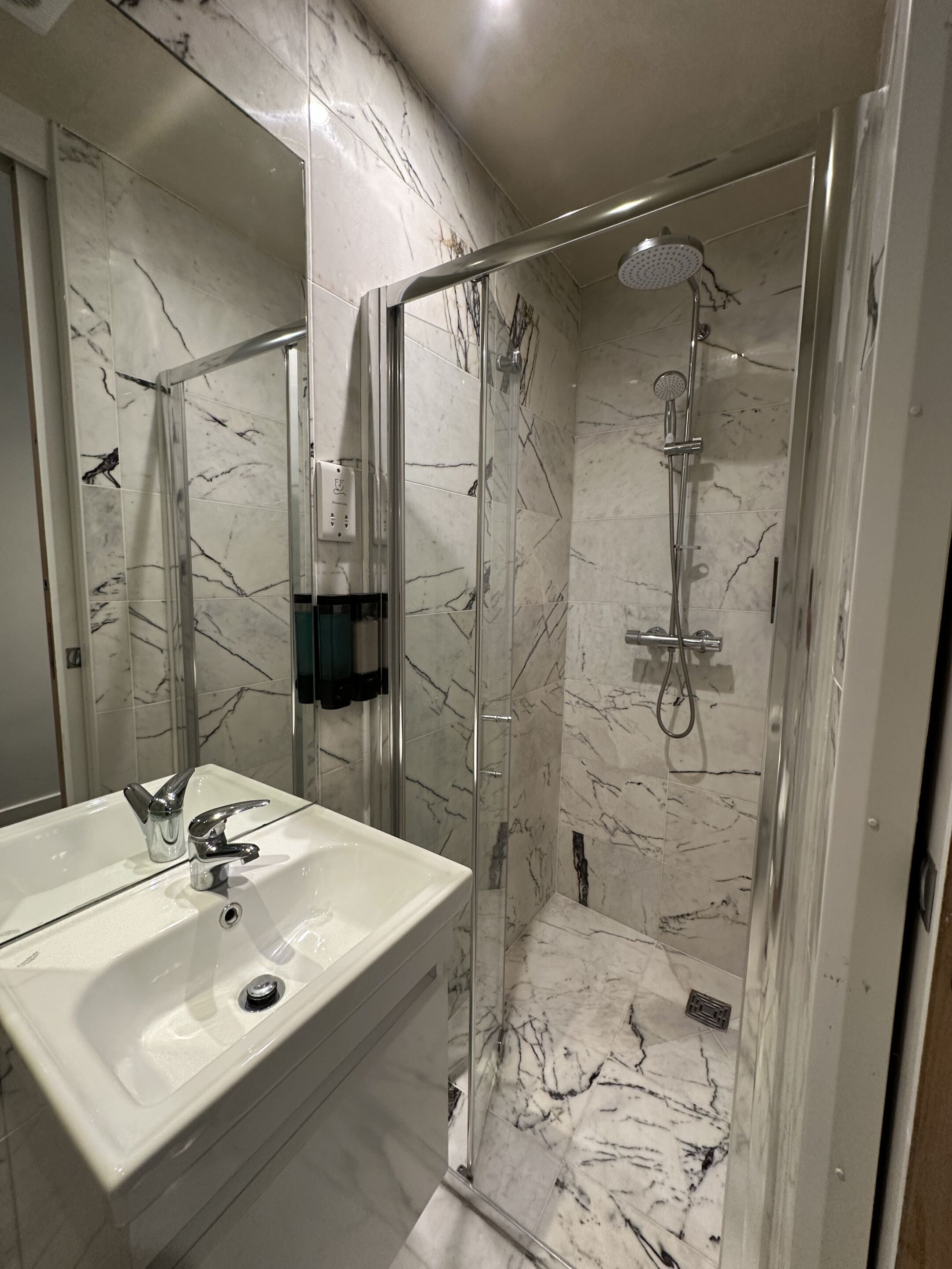 Un baño con ducha de vidrio, un lavamanos blanco con grifo cromado y paredes con diseño de mármol.