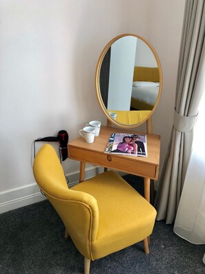 Una habitación compacta con un sillón amarillo, un pequeño escritorio de madera con un espejo, una revista y un secador de pelo.