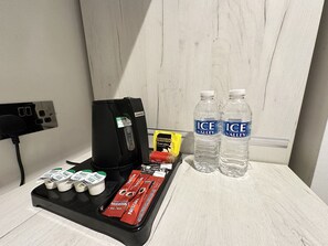Un minibar de hotel con una cafetera, botellas de agua y diferentes aperitivos.