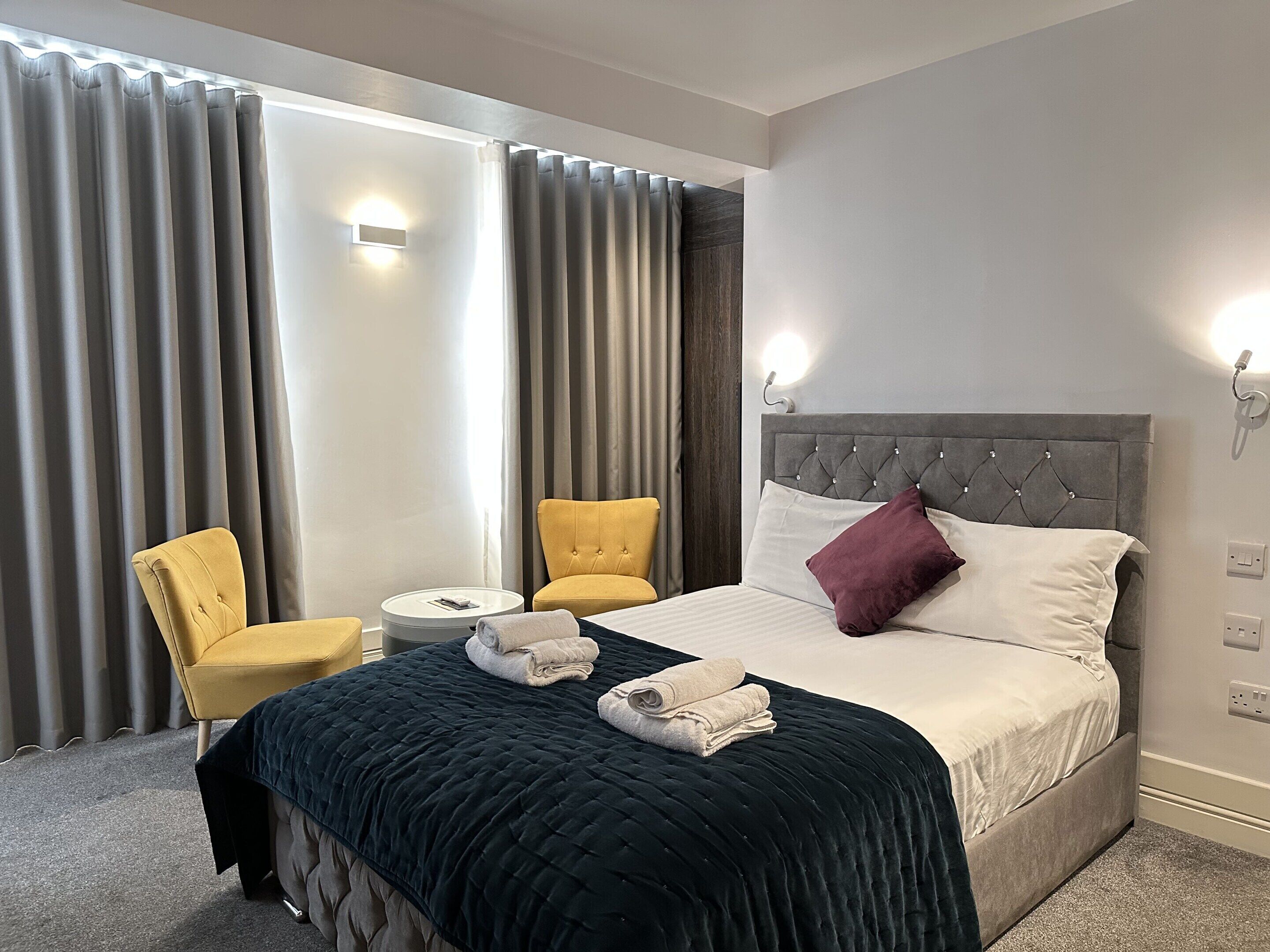 Habitación de hotel con cama, dos sillones, una mesa redonda y un ventanal con cortinas.