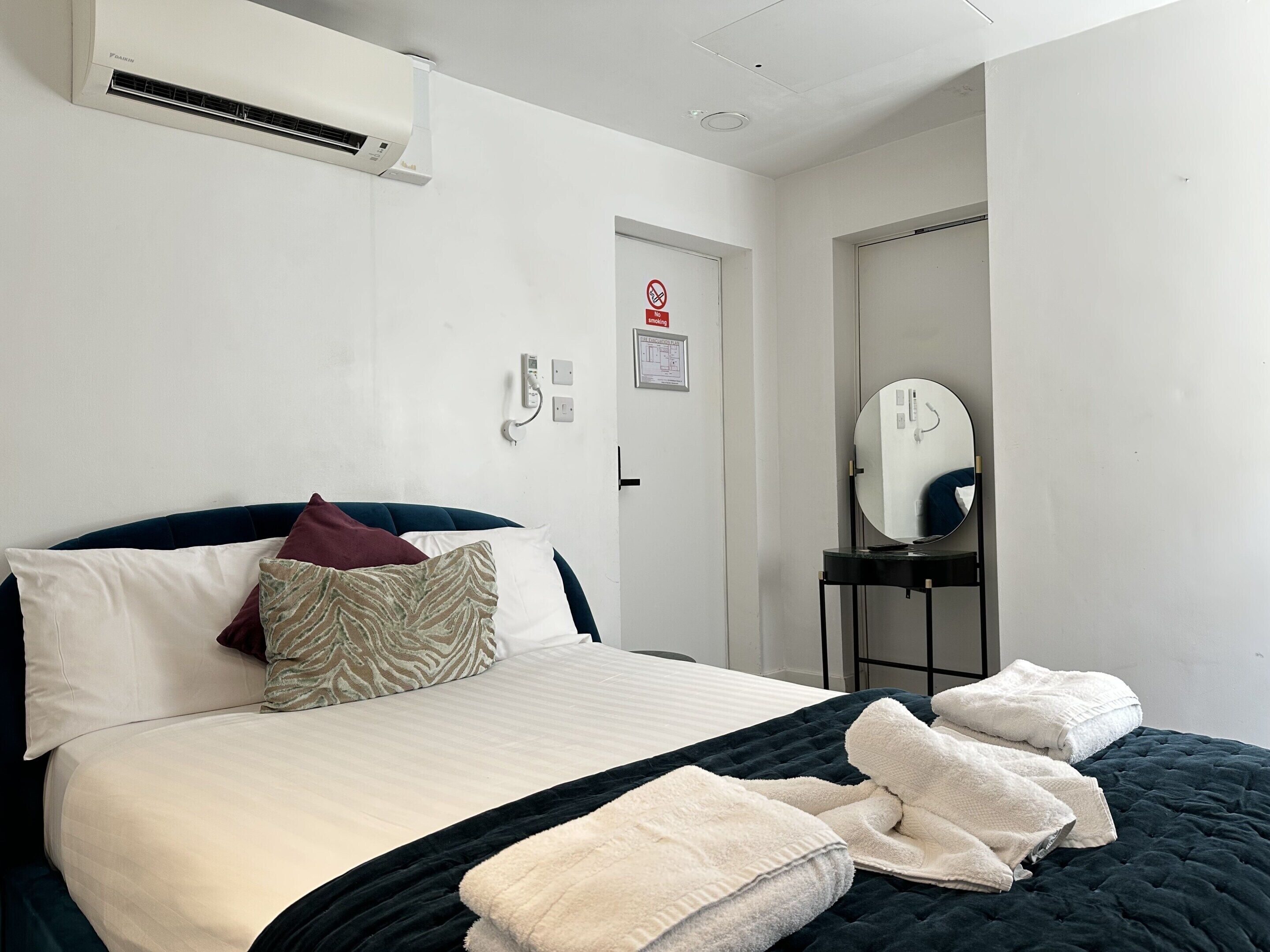 A hotel room with a bed, a bedside table with a mirror, a wall-mounted air conditioner, and a no-smoking sign.