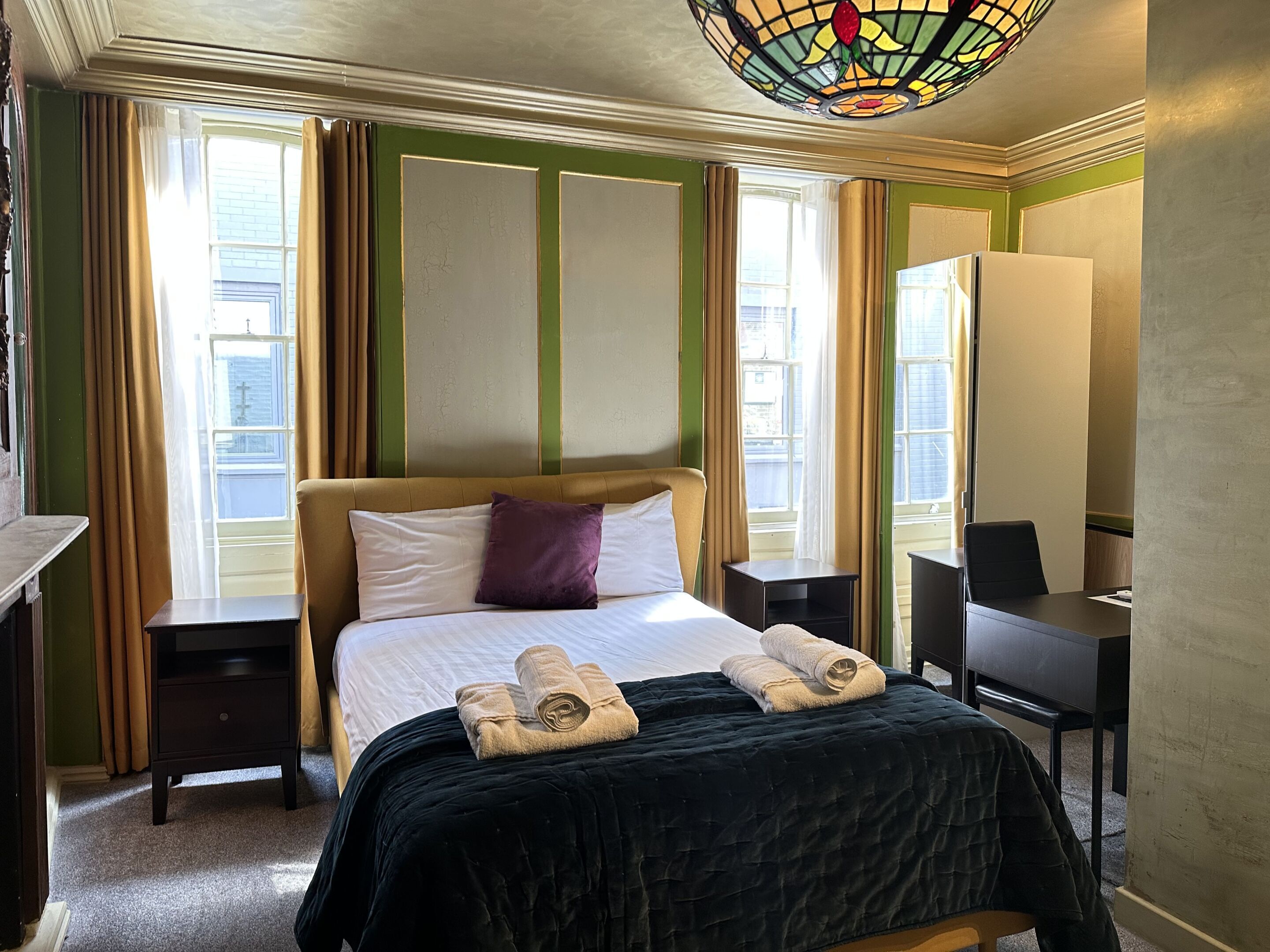 A hotel room with a bed, a desk, a chair, a small table, and a stained glass light fixture.