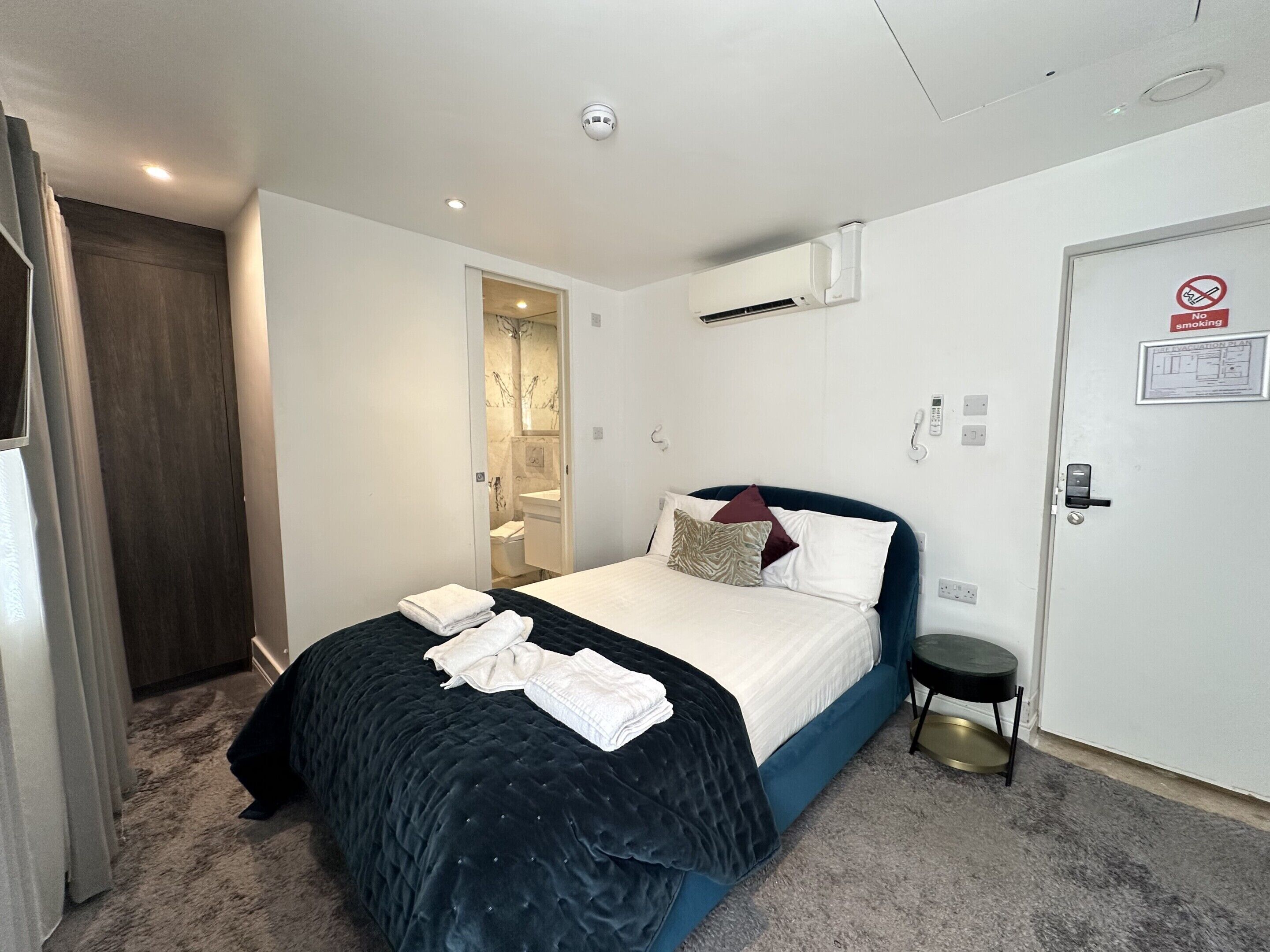 Quarto de hotel com uma cama, mesa de cabeceira, porta com placa de 'proibido fumar' e aparelho de ar condicionado.