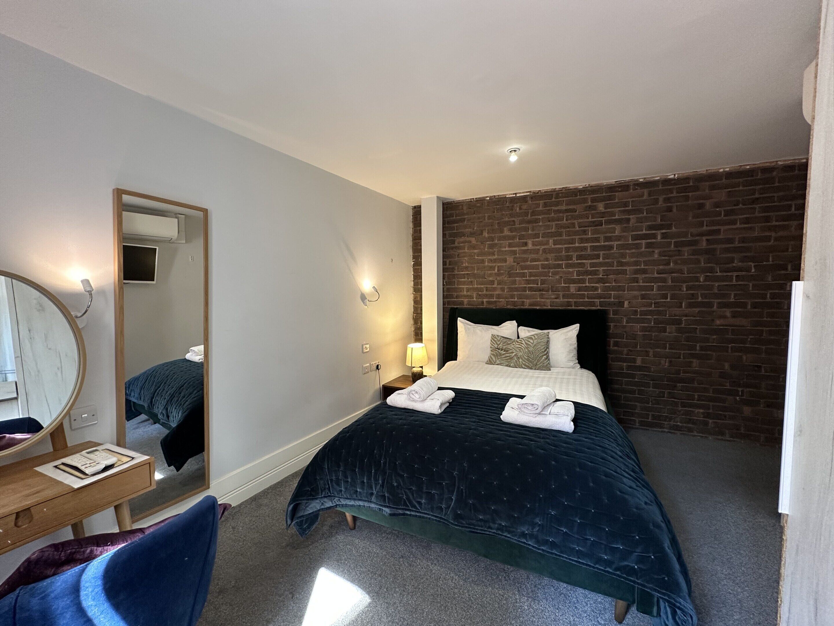 A hotel room with a bed, a desk, a chair, a mirror, and a brick wall.