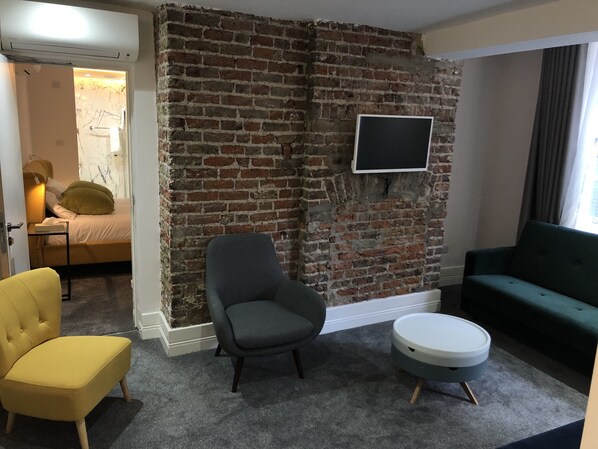 Une pièce avec un mur en briques, un lit, un fauteuil gris, un fauteuil jaune, un canapé vert et une petite table ronde.