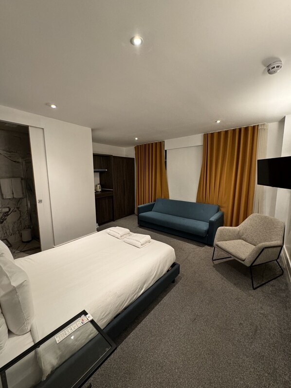 A hotel room with a bed, a blue sofa, a chair, a TV, and a wardrobe. - Widegate Residential (London)
