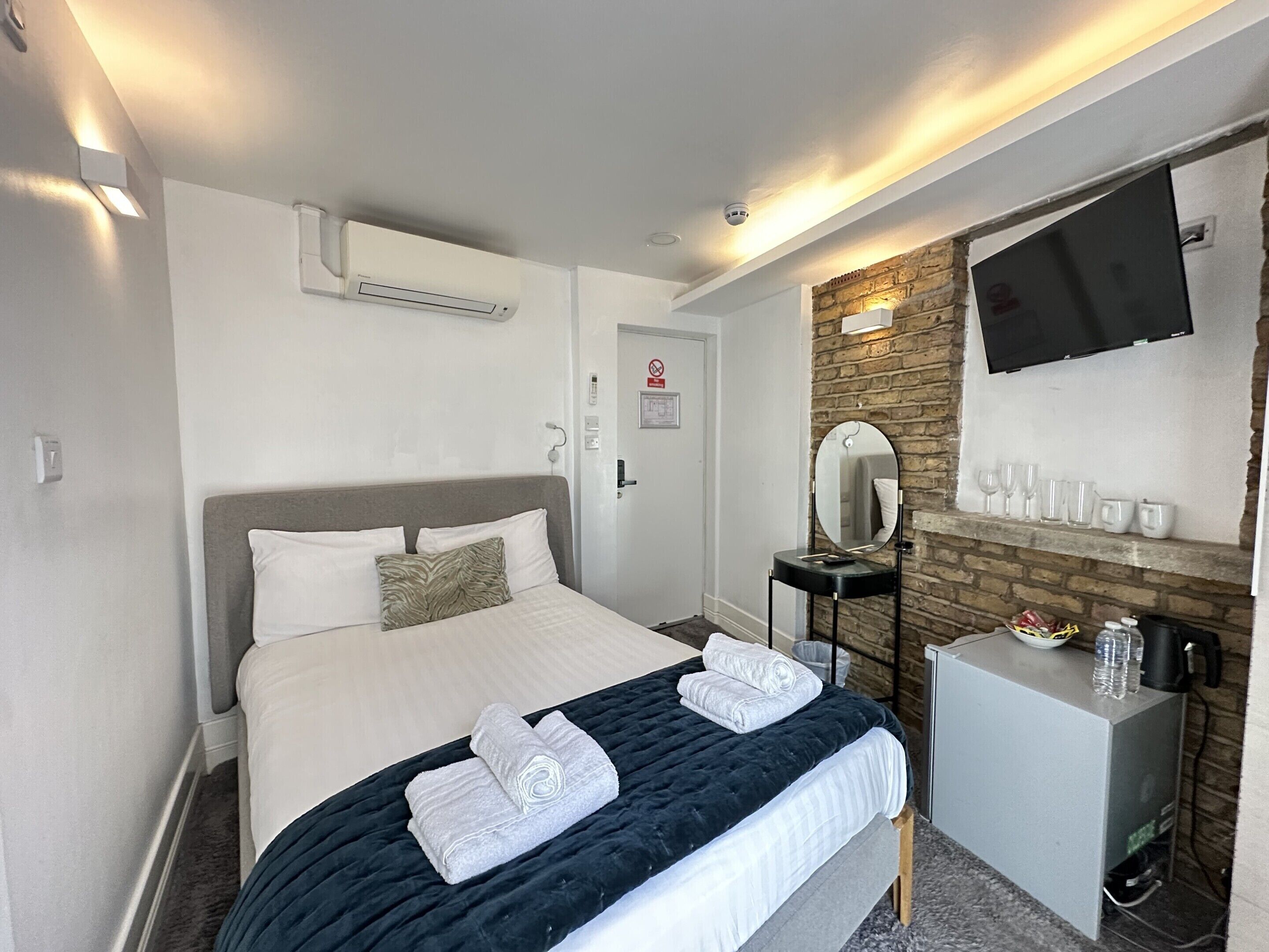 A hotel room with a bed, a bedside table, a mirror, a TV, and a wall-mounted shelf with glasses.