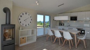 Une cuisine moderne avec des meubles blancs, un coin repas avec des chaises blanches, une grande horloge murale et un poêle à bois.