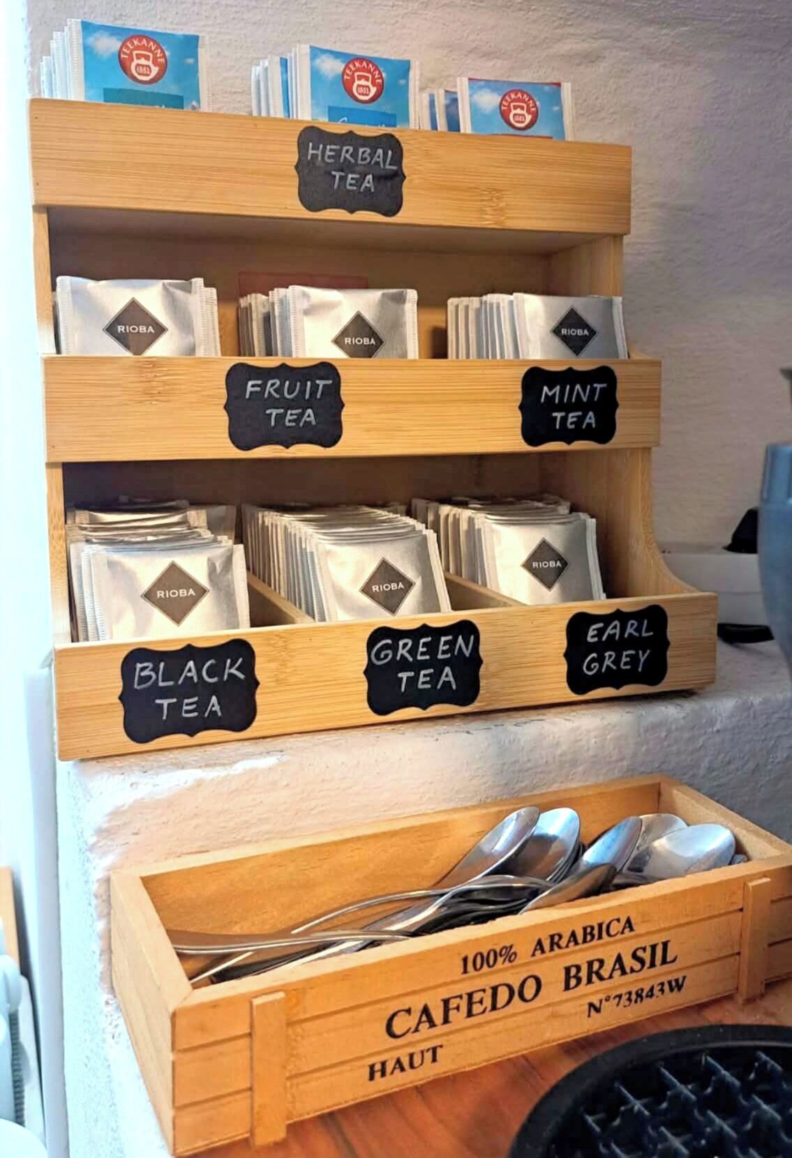 Een presentatie van verschillende soorten thee, zoals kruiden-, fruit-, mun-, zwarte, groene en Earl Grey-thee, met een houten kist met de tekst '100% Arabica Cafedo Brasil' onderaan.