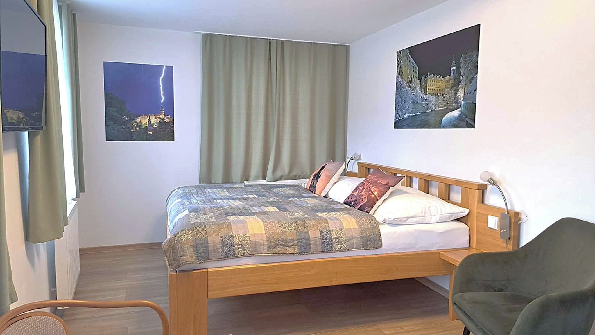 Een hotelkamer met een houten bed, een stoel, een raam met gordijnen en twee aan de muur bevestigde foto's.