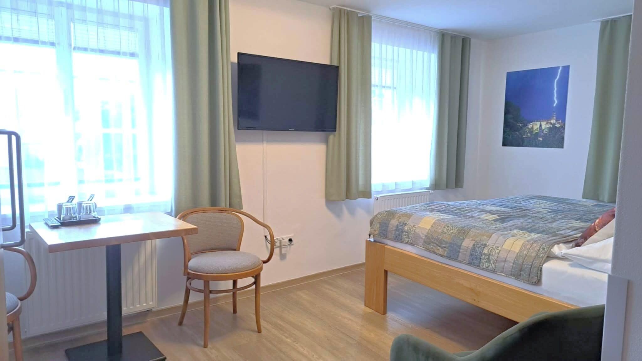 Habitación de hotel con cama, escritorio, silla, televisor y ventana con cortinas.