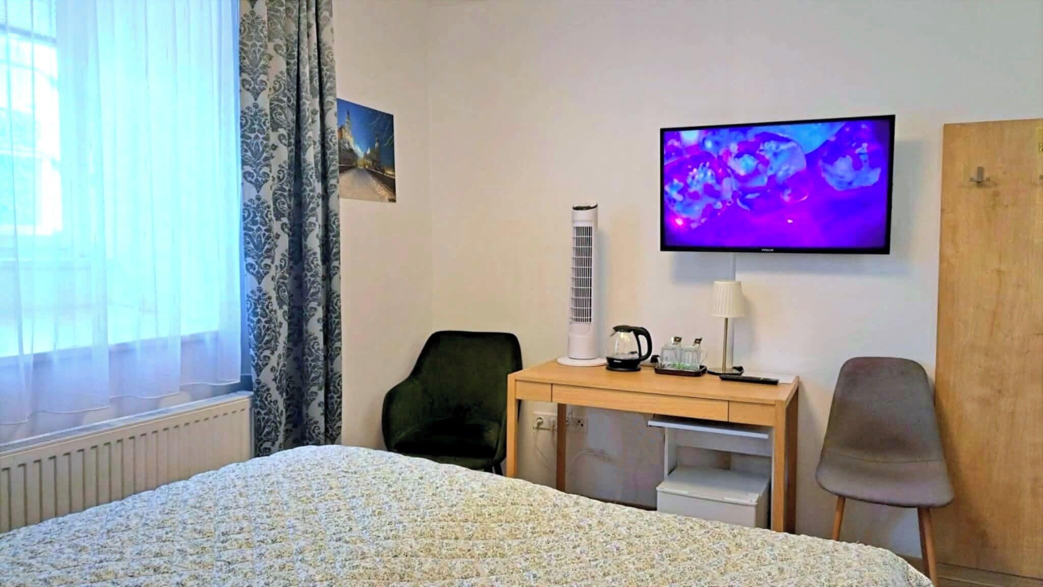 A hotel room with a bed, a desk with a lamp, a chair, a television, and a radiator.