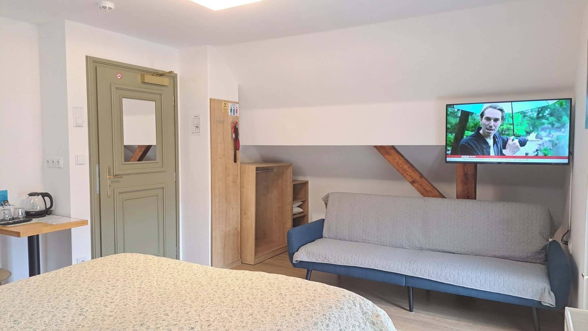 Una habitación con una cama, un televisor fijado a la pared, un armario de madera y una mesita con una tetera.