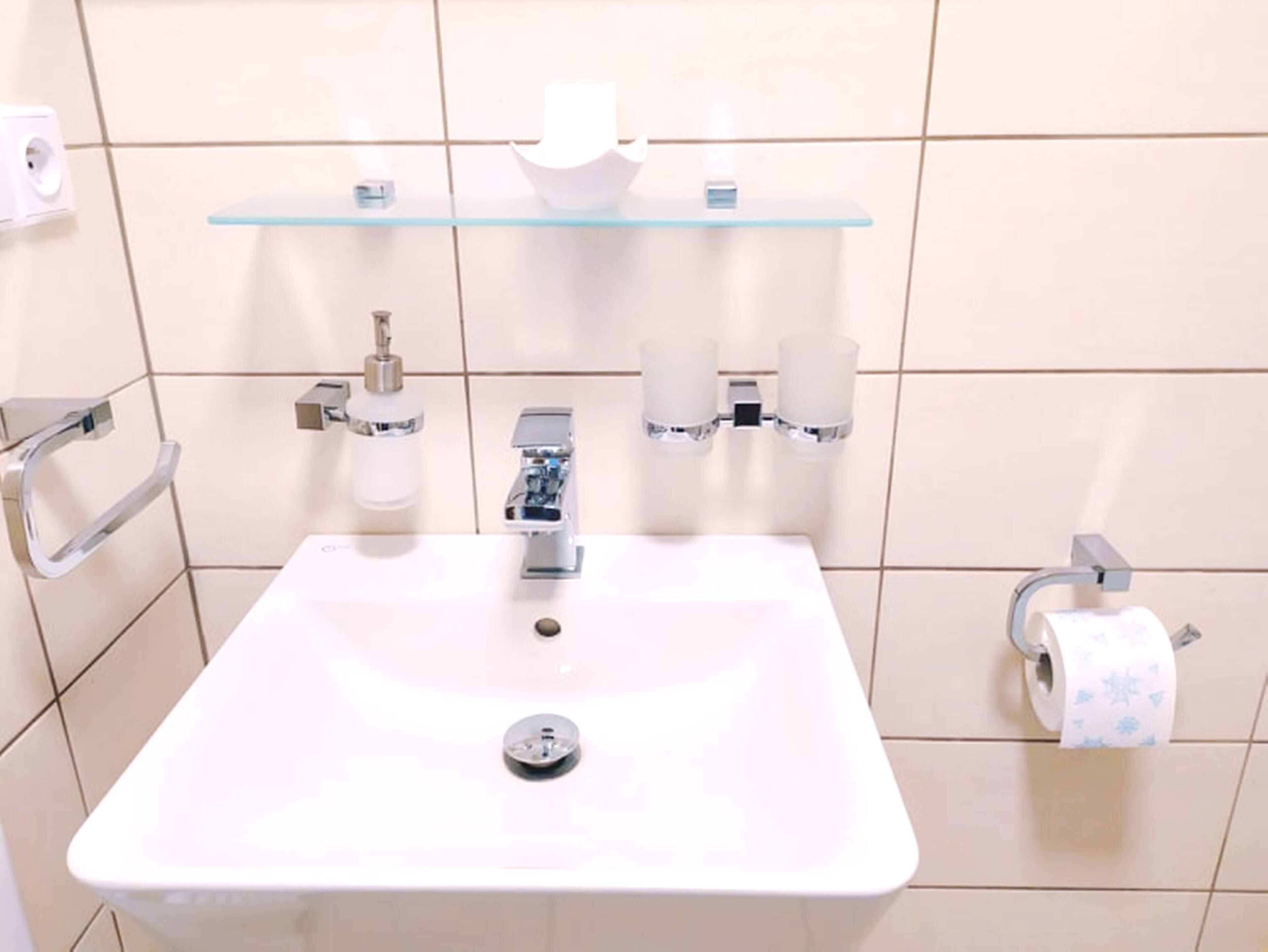 Un baño con un lavabo blanco, grifos cromados, un estante de vidrio sobre el lavabo y un dispensador de papel higiénico a la derecha.