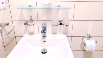 Un baño con un lavabo blanco, grifos cromados, un estante de vidrio sobre el lavabo y un dispensador de papel higiénico a la derecha.