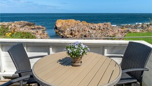 Un tavolo rotondo con piano color beige, circondato da due sedie in vimini, su una terrazza affacciata su una costa rocciosa e l'oceano.