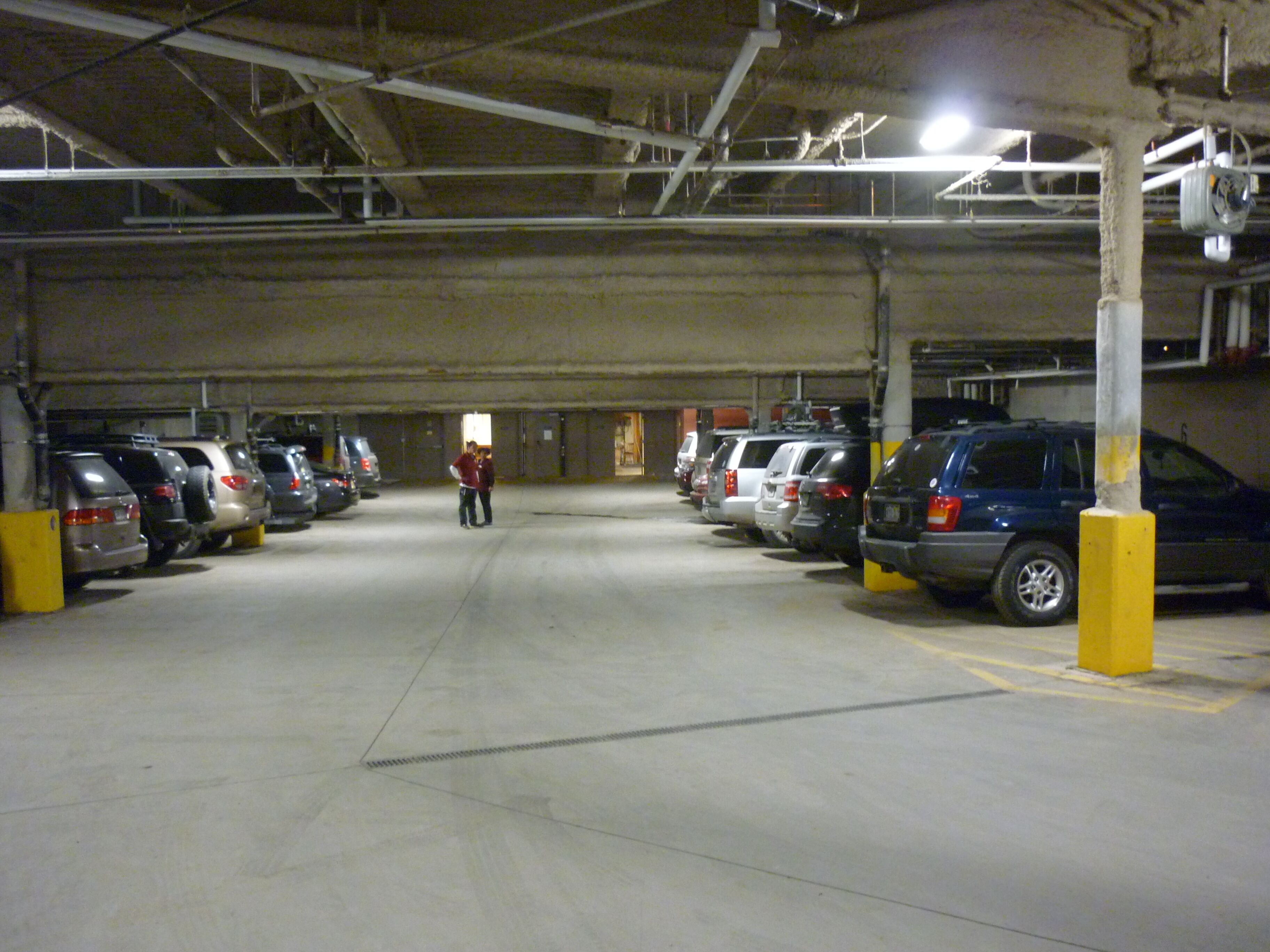 Un garaje de estacionamiento con varios coches aparcados en filas, pilares de hormigón e iluminación en el techo.