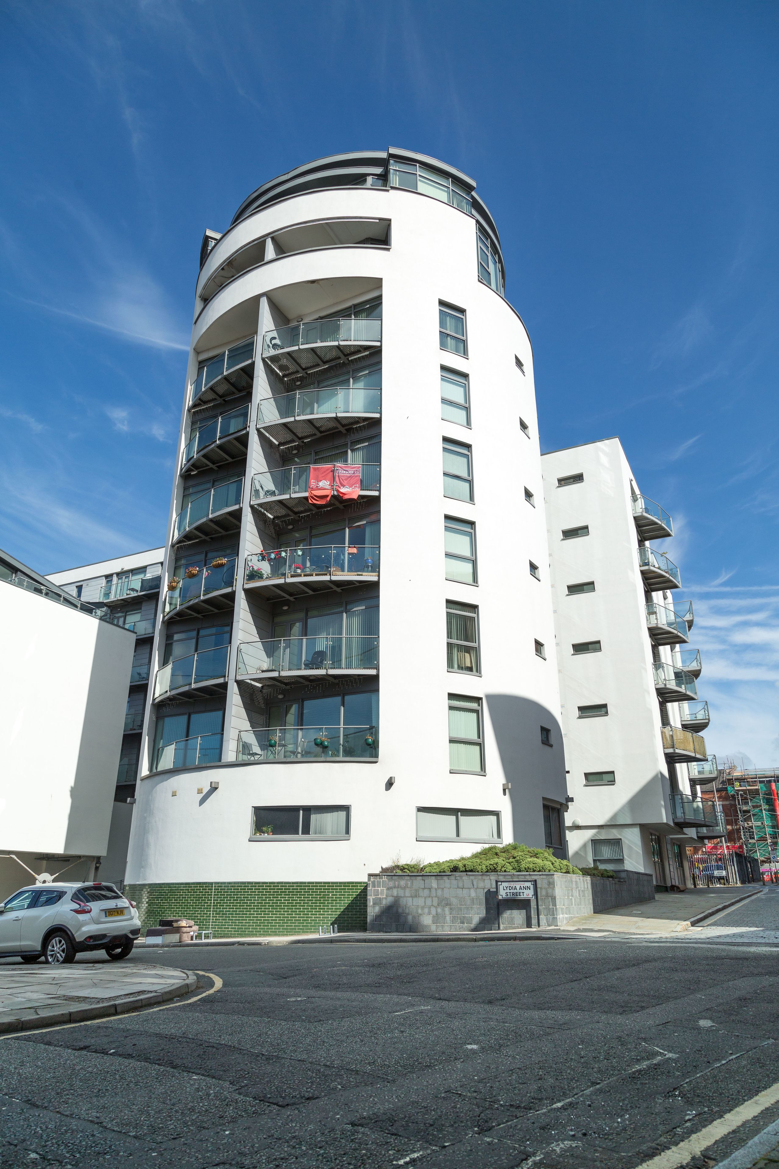 Un immeuble moderne à plusieurs étages, doté de balcons, avec une voiture garée devant et un ciel bleu dégagé.
