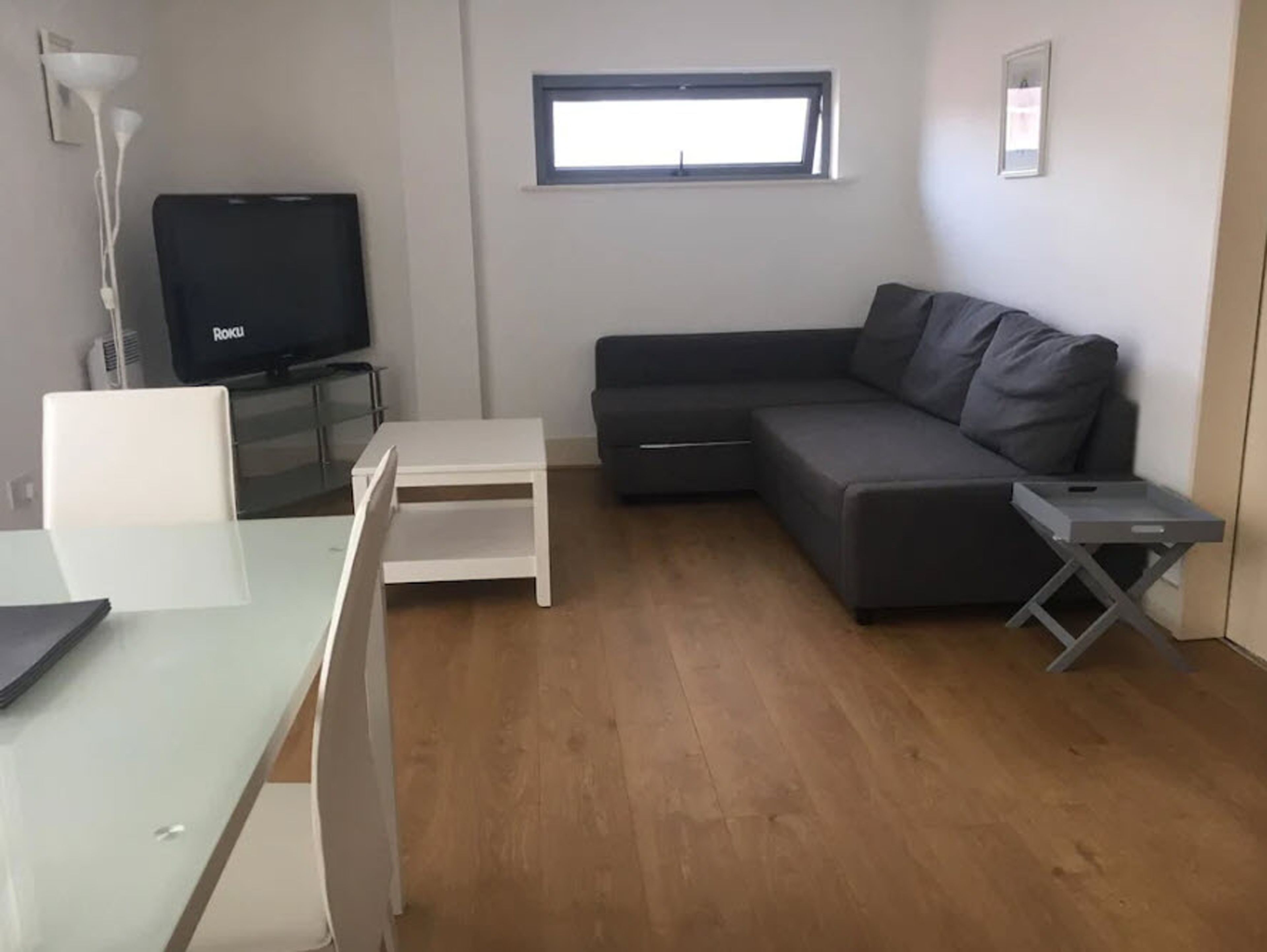 A modern living room with a grey sofa, a white coffee table, a TV on a stand, and a small window.