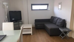 Uma sala de estar moderna com um sofá cinzento, uma mesa de centro branca, uma TV em um móvel e uma janela pequena.