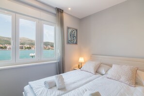A hotel room with a bed, a nightstand, a lamp, a painting, and a window with a view of a body of water and buildings. - 4 Elements Old Town Views Rooms (Trogir)