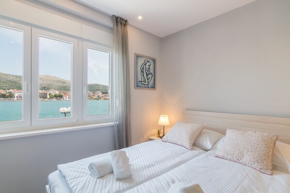 Una habitación de hotel con cama, mesita de noche, lámpara, cuadro y ventana con vista a un cuerpo de agua y edificios.