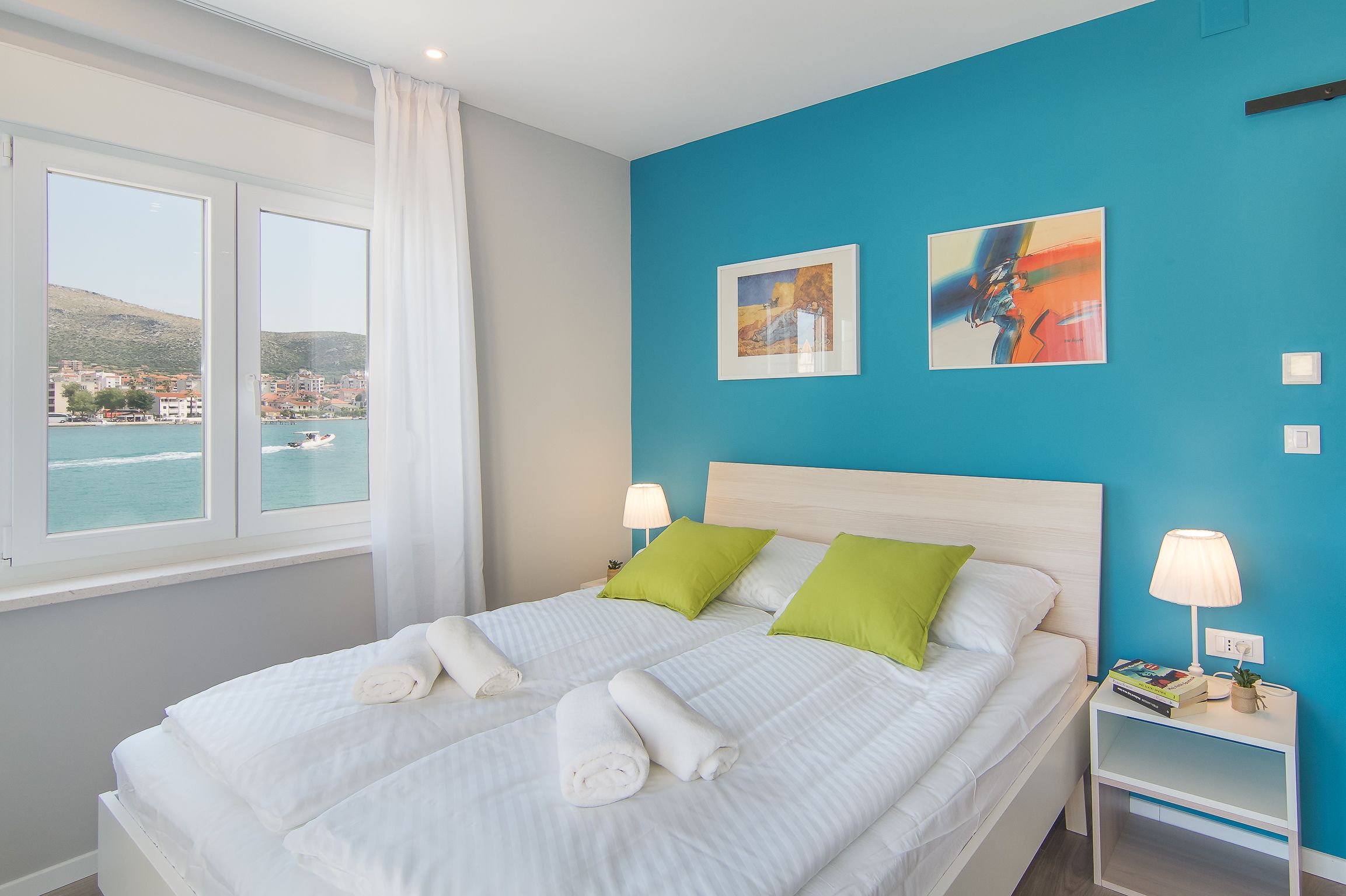 A double bed with white linens and green pillows, a nightstand with a lamp, and two framed artworks on a blue wall.