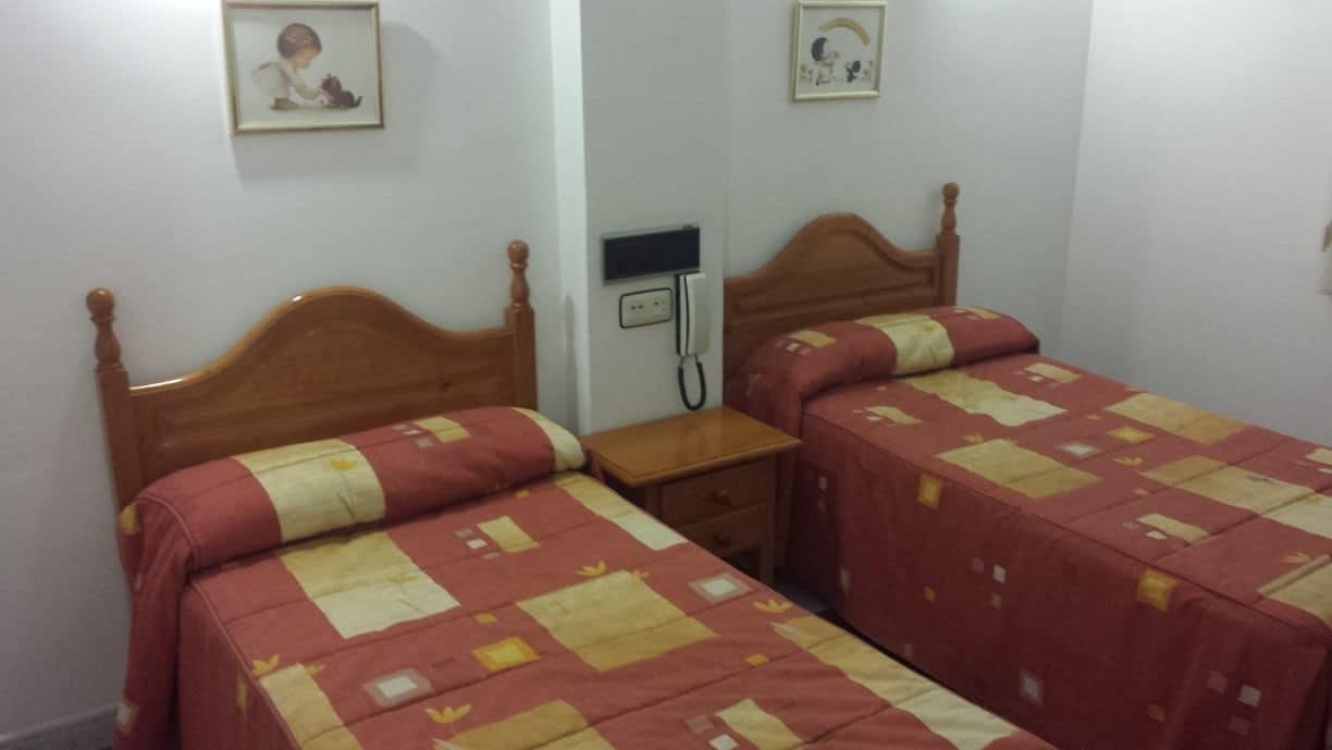 A room with two wooden beds, each with a patterned bedspread, a bedside table, and a wall-mounted telephone.