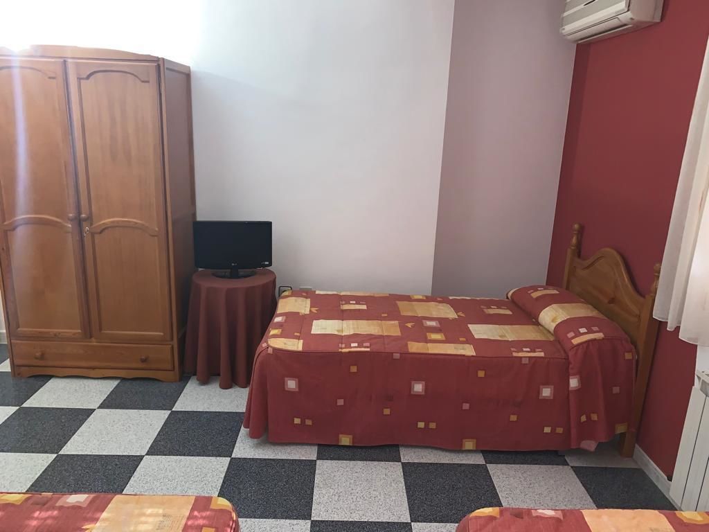 Une chambre avec un sol à carreaux, un lit recouvert d’une couvre-lit à motifs rouges et jaunes, une armoire en bois et une petite table ronde sur laquelle se trouve un téléviseur.