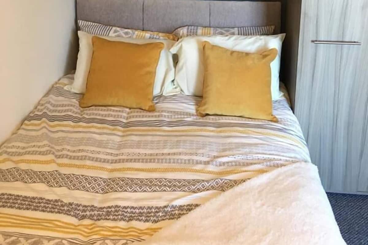 A neatly made bed with patterned bedding, a grey headboard, and two pillows. Above the bed, there is a shelf with a framed artwork and a small table lamp. To the right, there is a wardrobe with a light wood finish.