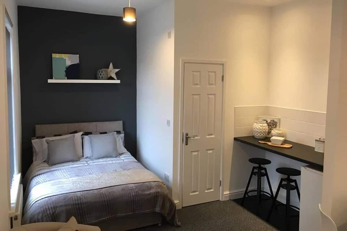 A bedroom with a bed, a nightstand, a desk with stools, and a door.