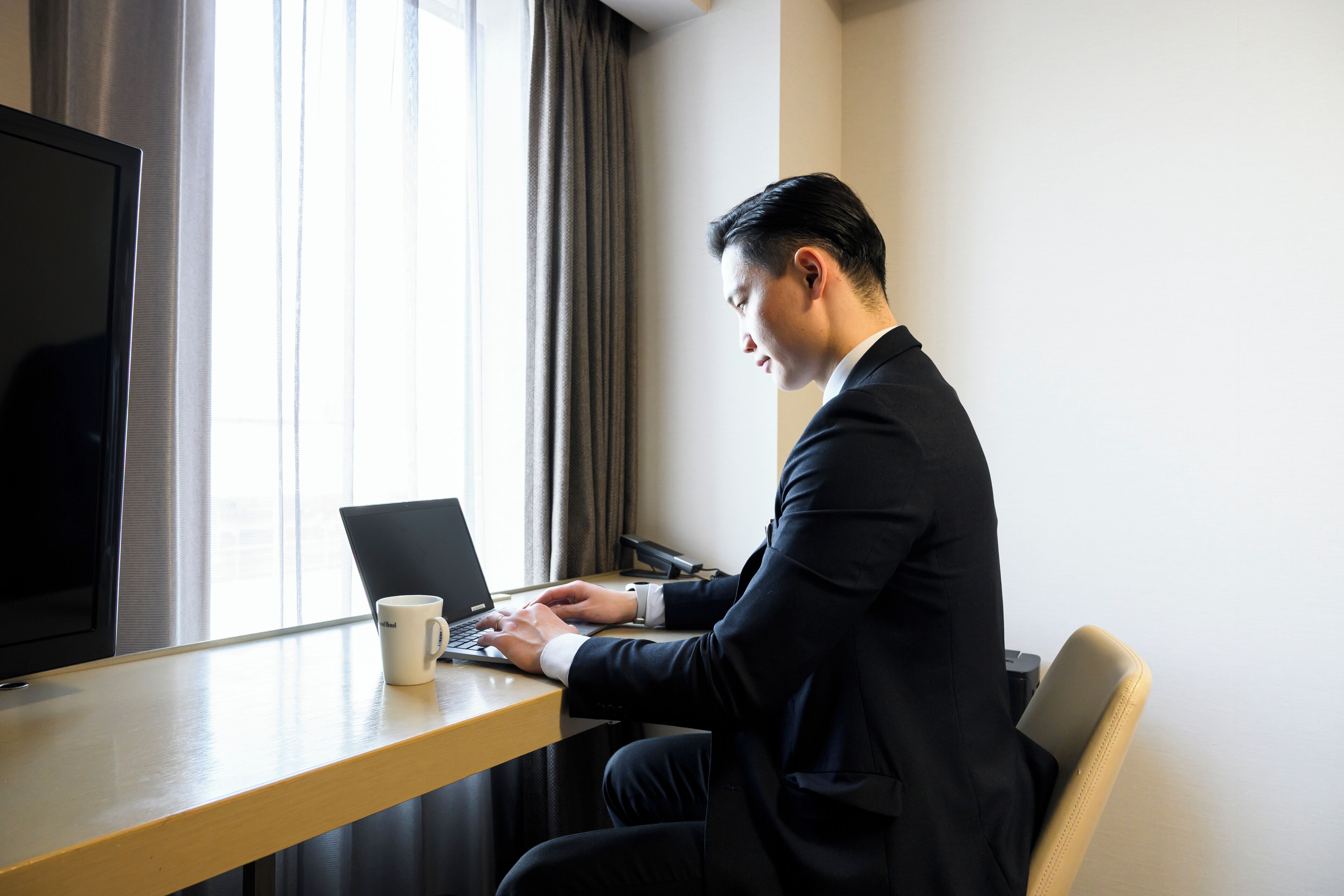 Un hombre con traje trabaja en un escritorio con una computadora portátil, una taza de café y un televisor en una habitación con ventana y cortinas.