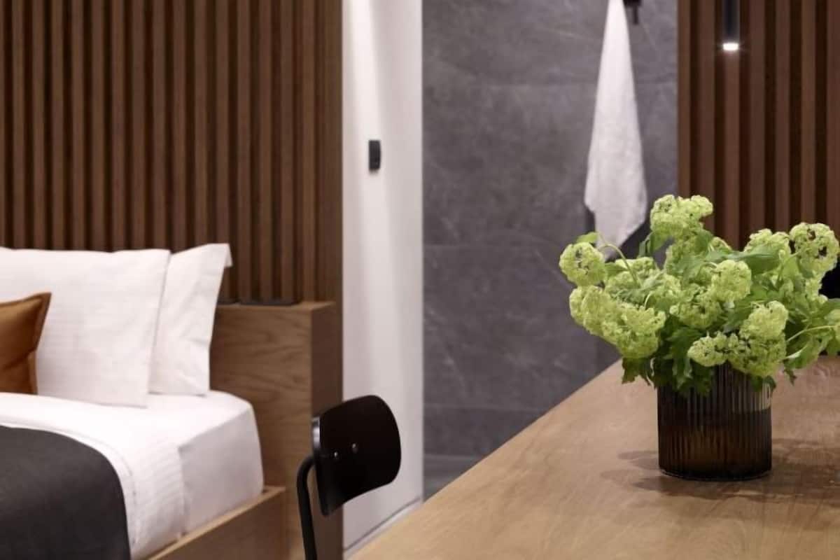 Habitación de hotel moderna con una cama de madera, una mesita con una planta verde y vistas a la ciudad a través de amplios ventanales.