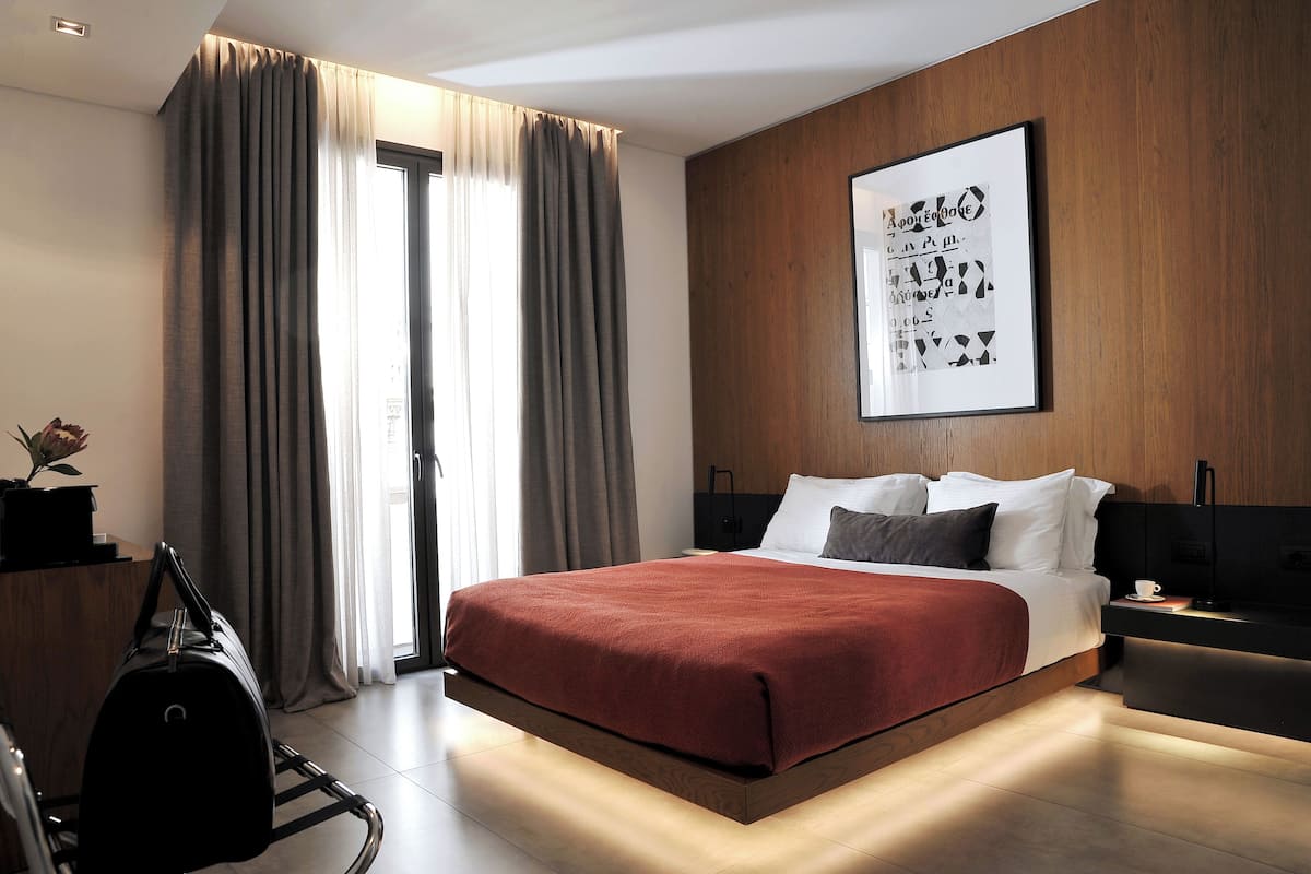 Una habitación de hotel moderna con una cama grande, un cabecero de madera, una mesita de noche con lámpara y un ventanal con cortinas.