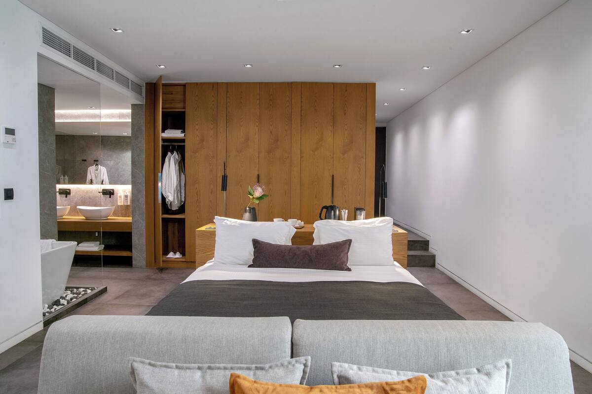 Una habitación de hotel moderna con una cama grande, un armario de madera, un baño con espejo y un pequeño rincón de descanso.